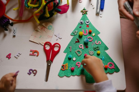 A child's hands creating a festive Christmas tree craft using colorful decorations, scissors, and craft supplies on a table.